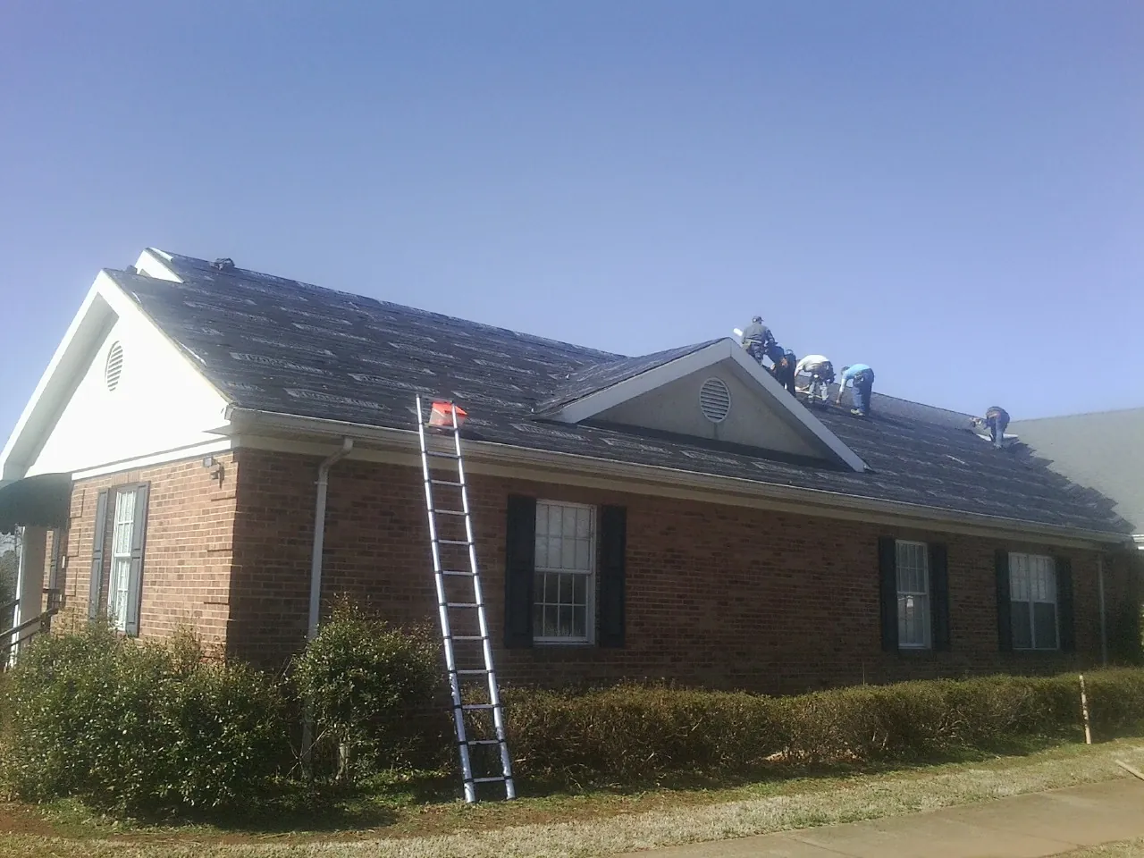 Crew tearing shingles off a brick ranch roof with a ladder set — Emergency Roof Repair work by our crew in Primghar