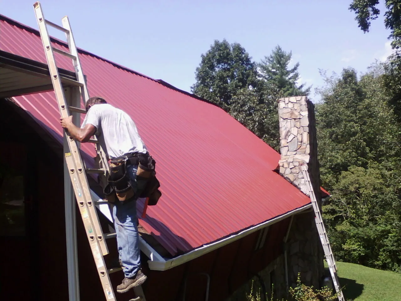 Roofer on a ladder beside a red metal roof and stone chimney — Soffit & Fascia Repair by Roofing Brothers in Primghar